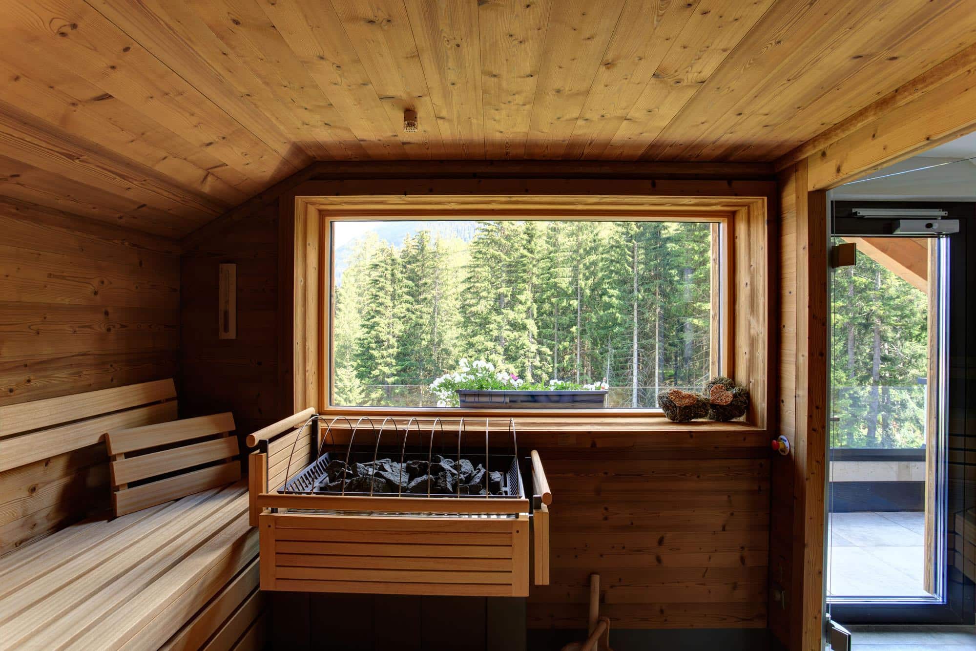Die Holzsauna im 4 Sterne Hotel St. Anton besticht durch ein großes Fenster mit herrlichem Waldblick. Bänke säumen die Wände, während ein Steinofen im Vordergrund Wärme und Entspannung verspricht.