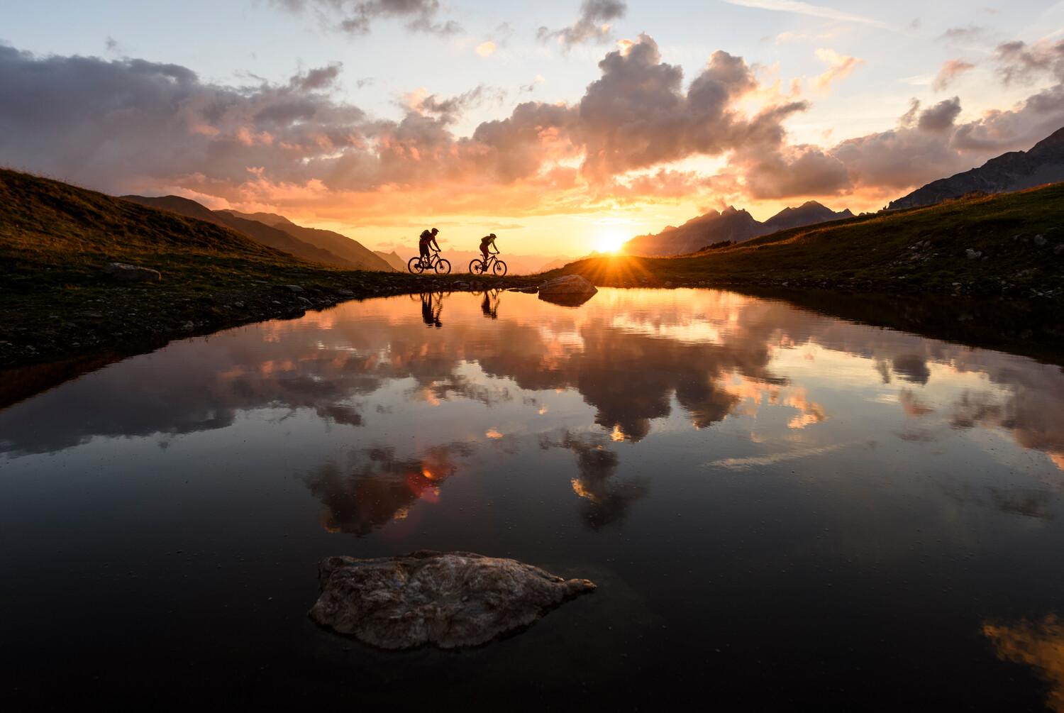Zwei Menschen fahren bei Sonnenuntergang mit dem Fahrrad neben einem spiegelnden See entlang, mit Bergen im Hintergrund und einem dramatischen Himmel.