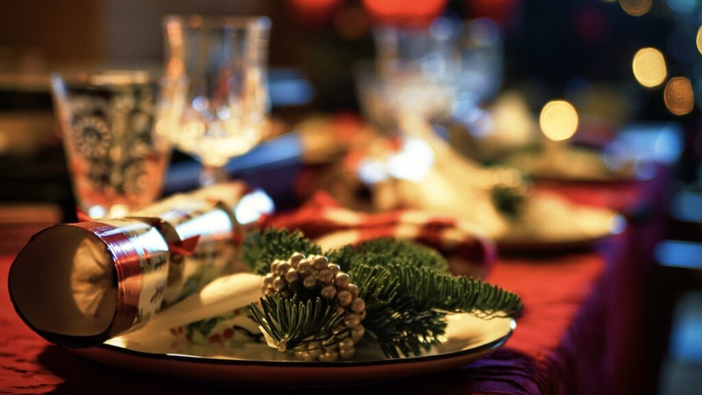 A festive table setting with glassware, plates, and holiday greenery on a red tablecloth.