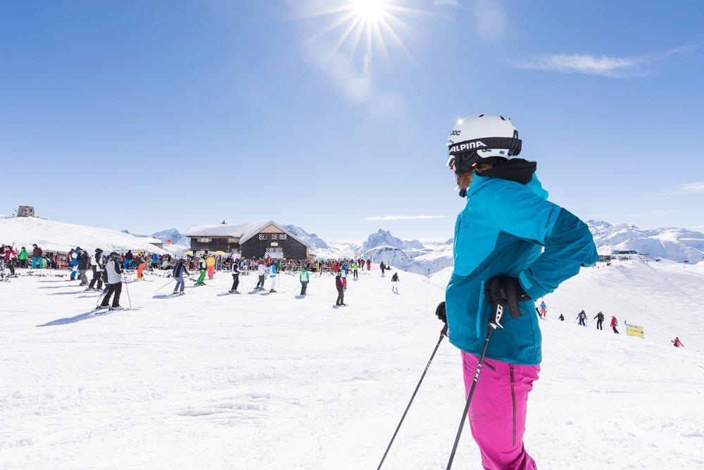 Skifahrer in blauer Jacke und rosa Hose steht auf einer verschneiten Piste unter sonnigem Himmel.