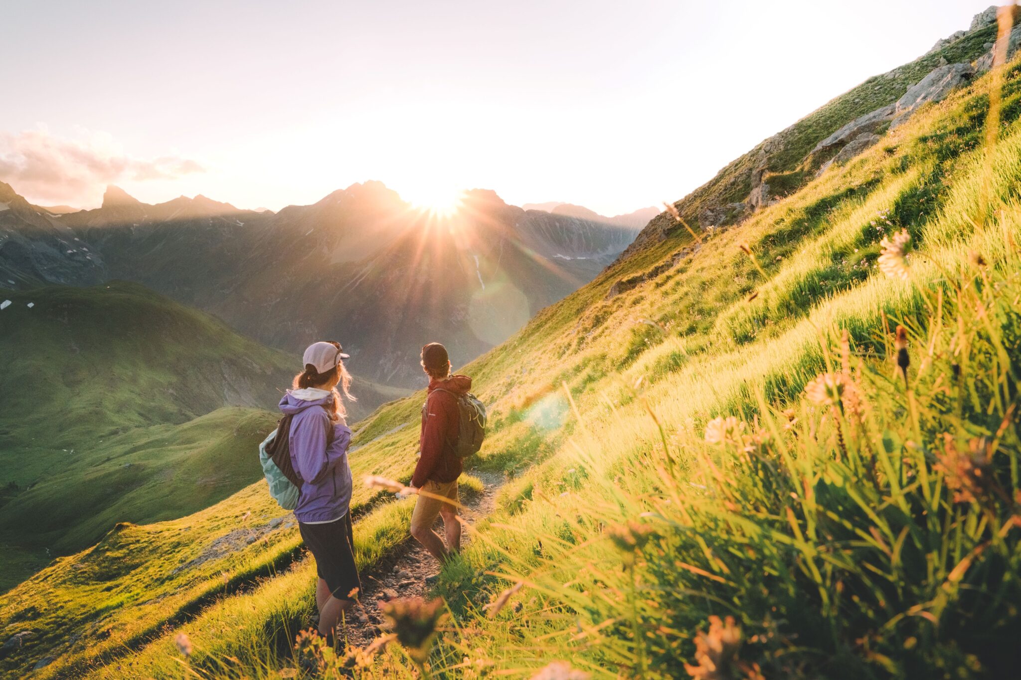 Zwei Wanderer gehen bei Sonnenaufgang auf einem grasbewachsenen Bergpfad mit Sonnenstrahlen über den fernen Gipfeln.