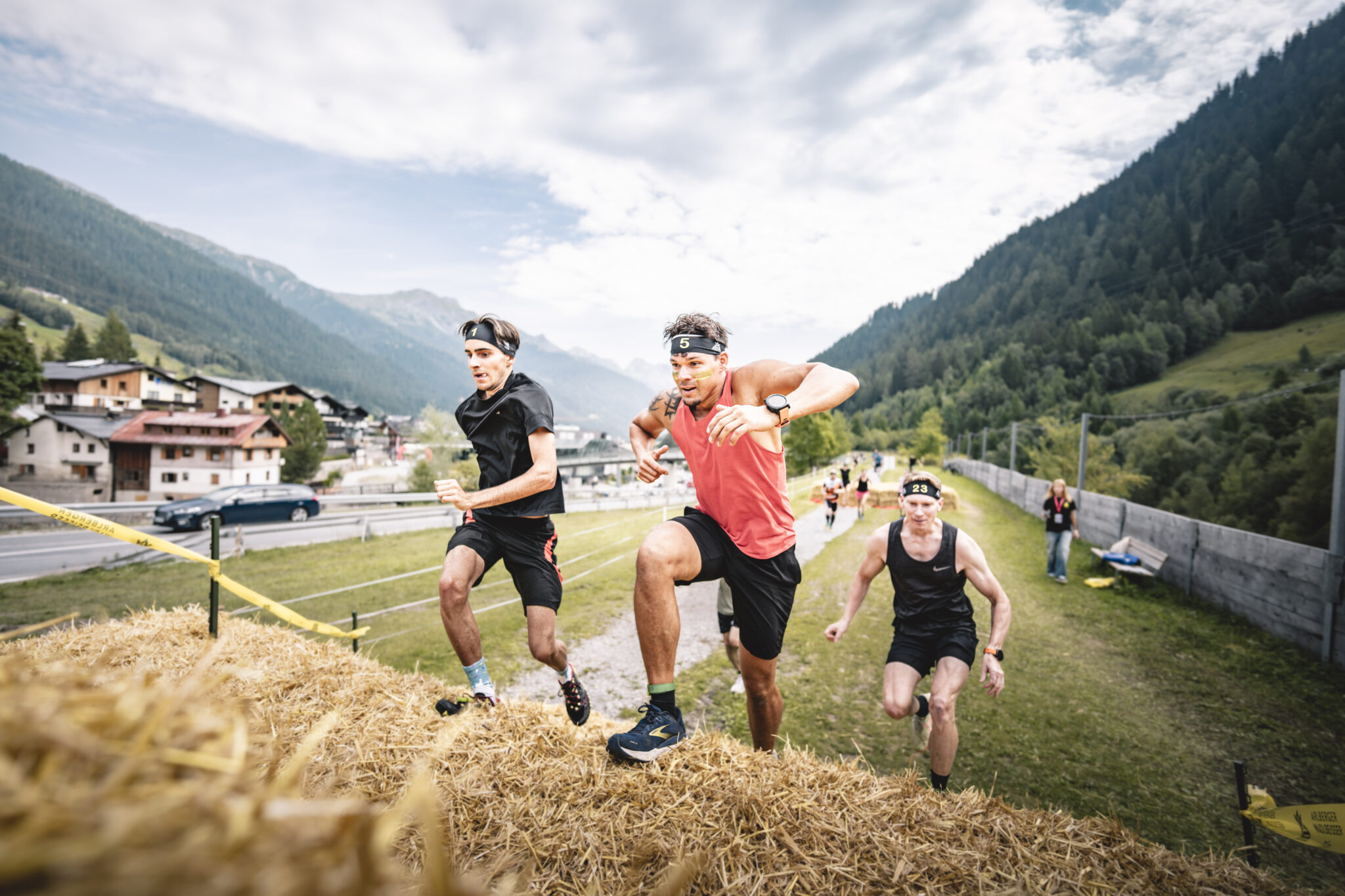Three men in athletic gear run up a hay bale obstacle in an outdoor race with mountains behind.