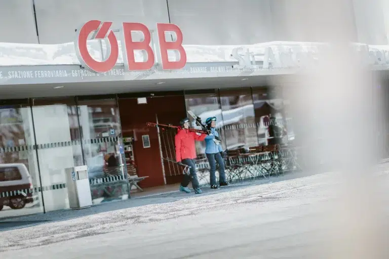 Zwei Personen mit Skiern gehen vor einer ÖBB-Station beim Hotel an der Piste St. Anton im Schnee spazieren.